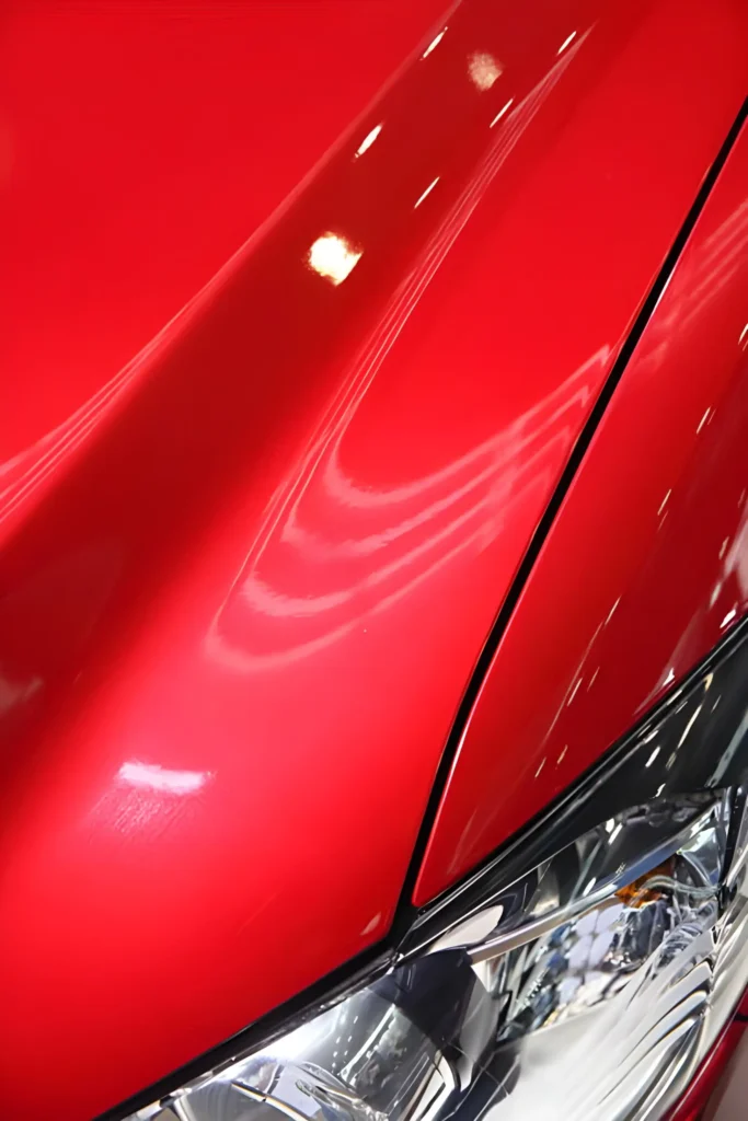 Close-up of a vibrant red car hood and headlight, showcasing brilliant, deep gloss paint reflection.